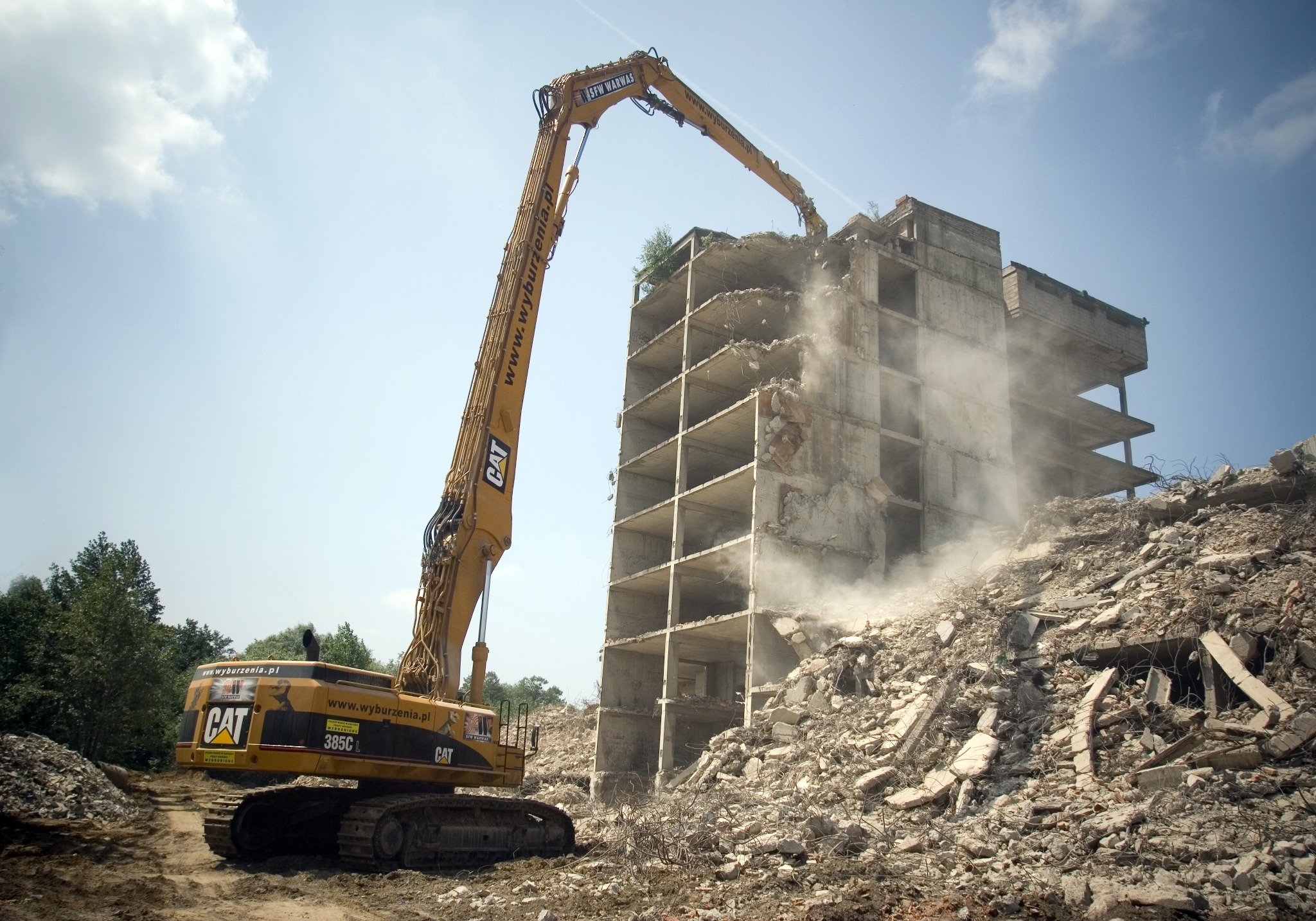 Żółty, wysięgnikowy dźwig CAT 385C rozbiera betonowy budynek. Gruzowisko u podnóża, błękitne niebo w tle. Widoczne logo firmy na maszynie.
