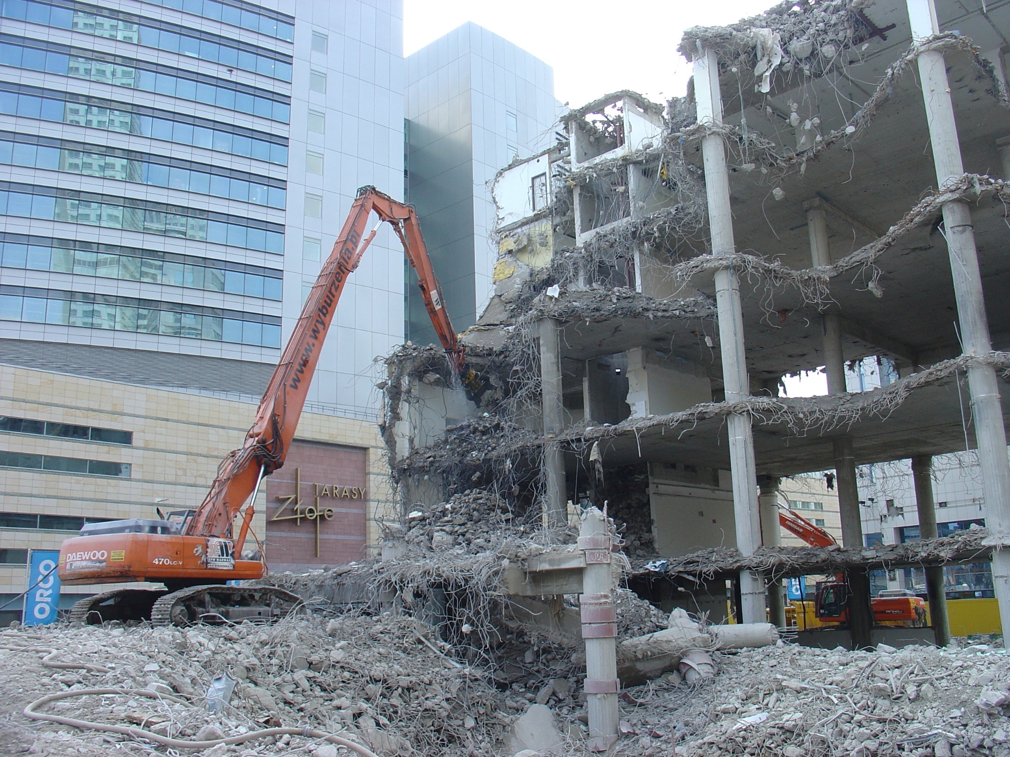 Pomarańczowy dźwig Daewoo 470LC-V rozbiera budynek Tarasy Złote, gruzowisko na pierwszym planie, w tle nowoczesna architektura.