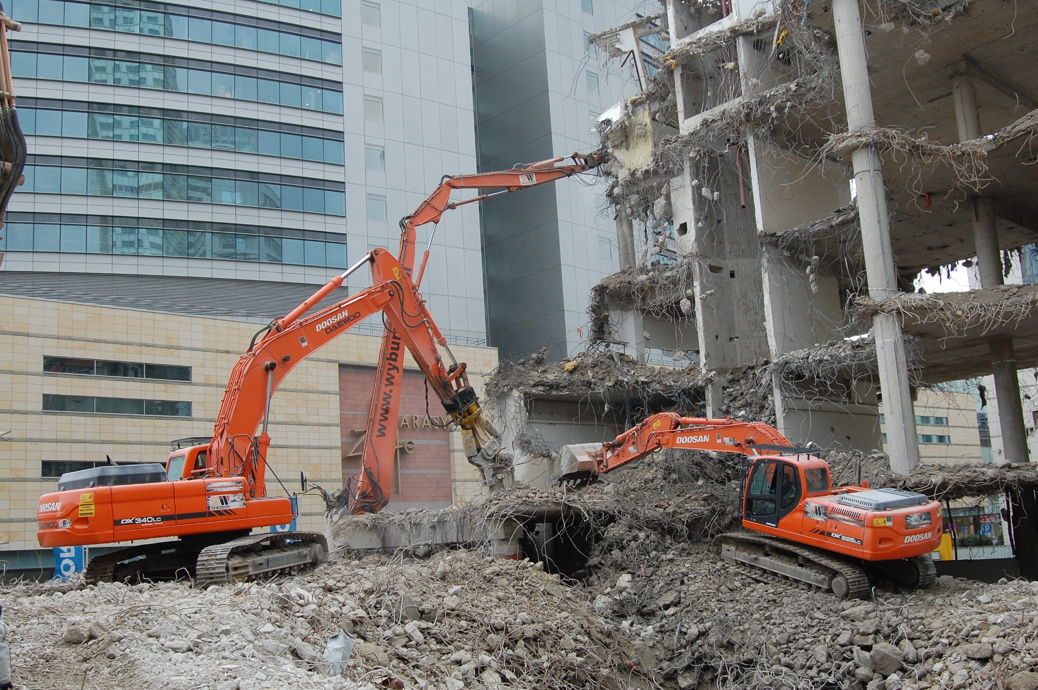 Pomarańczowe koparki Doosan na gruzowisku podczas rozbiórki budynku. W tle nowoczesny biurowiec z odbijającym się niebem w oknach.