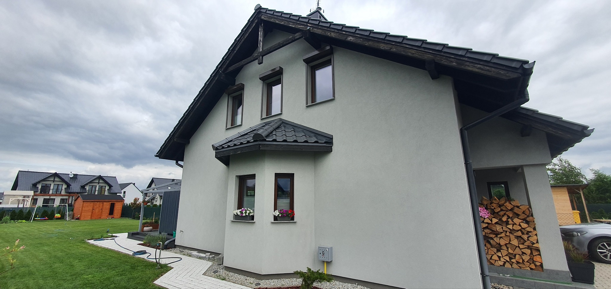 Dom z szarymi elewacjami i brązowymi oknami w Kamieńcu Ząbkowickim. Na oknach widoczne zwinięte osłony okienne. W oknach kwiaty w doniczkach.
