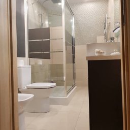 Stanisław Kach - Jasna łazienka z kabiną prysznicową, toaletą i bidetem. Umywalka z szafką w ciemnym kolorze. Na ścianie mozaika. Widok zza framugi drzwi.