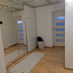 Stanisław Kach - Jasne wnętrze z lustrzaną szafą przesuwną, drewnianą podłogą i dywanikiem. Widoczne drzwi z przeszkleniami i kosz z poduszkami. Minimalistyczny design.
