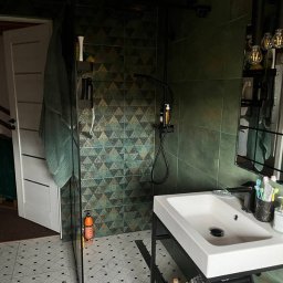 Stanisław Kach - Nowoczesna łazienka z zielonymi, geometrycznymi płytkami, czarną armaturą i białą umywalką na metalowym stelażu. Widoczny fragment kabiny prysznicowej i drewniany sufit.