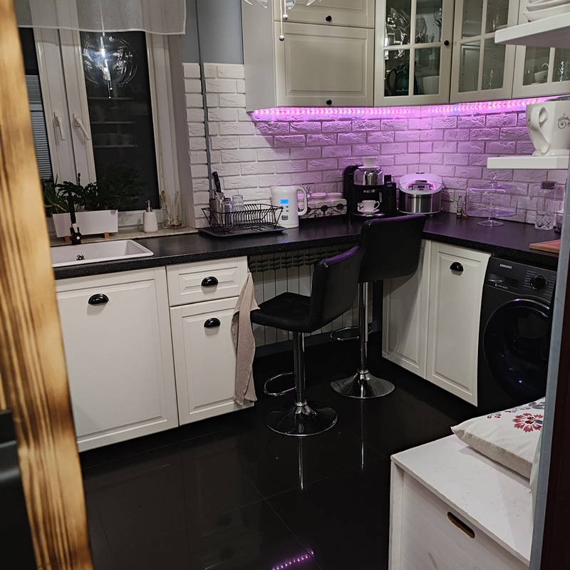 Biała kuchnia z czarnym blatem i podświetleniem LED. Dwa czarne hokery przy blacie. Na ścianie biała cegła. Błyszcząca, czarna podłoga. Nowoczesny design.