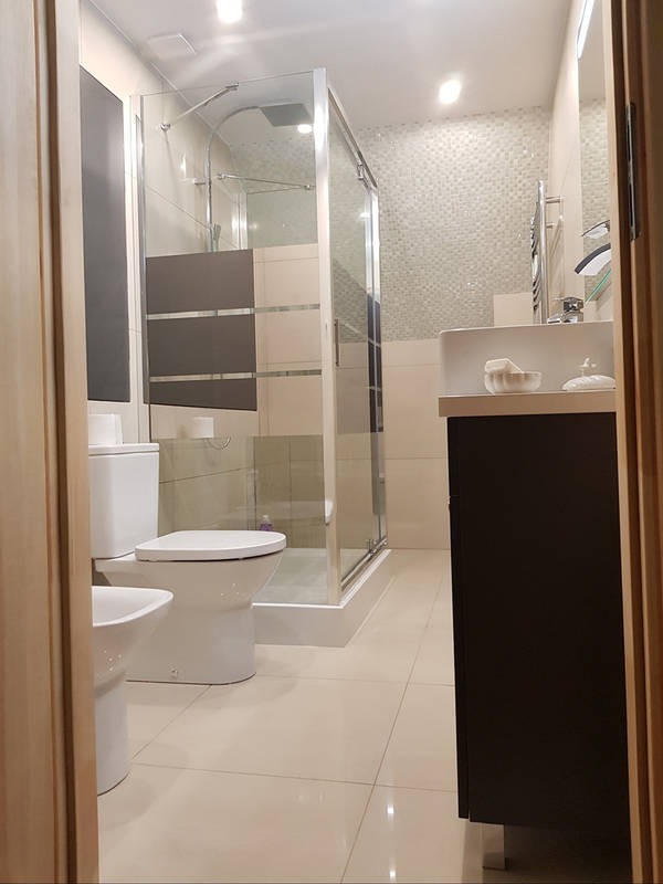 Jasna łazienka z kabiną prysznicową, toaletą i bidetem. Umywalka z szafką w ciemnym kolorze. Na ścianie mozaika. Widok zza framugi drzwi.