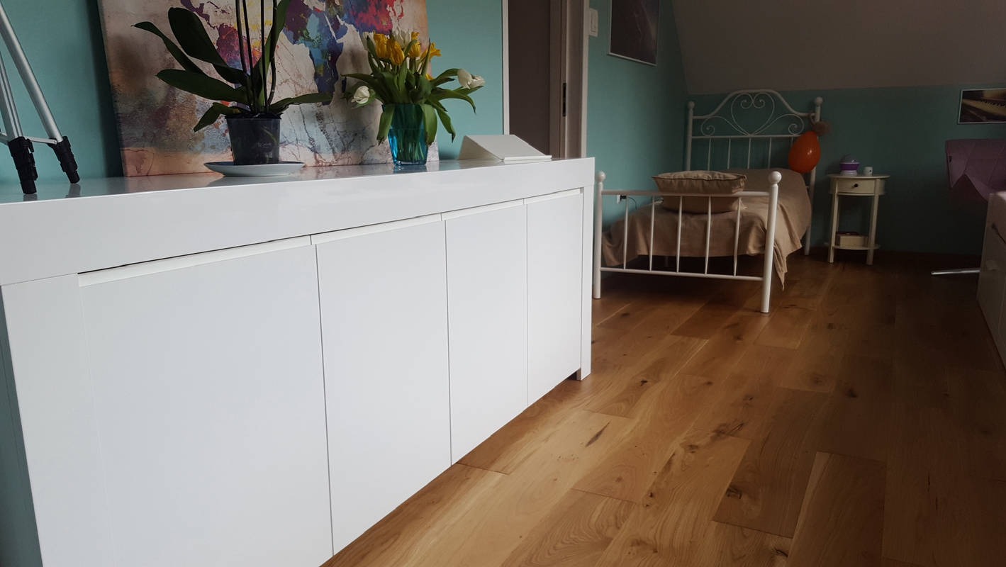 Jasne wnętrze pokoju z drewnianą podłogą, białymi meblami i turkusowymi ścianami. Na komodzie kwiaty i obraz. W tle białe łóżko z metalową ramą i stolik nocny.