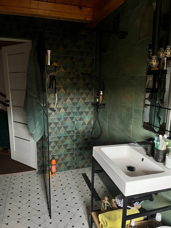 Nowoczesna łazienka z zielonymi, geometrycznymi płytkami, czarną armaturą i białą umywalką na metalowym stelażu. Widoczny fragment kabiny prysznicowej i drewniany sufit.
