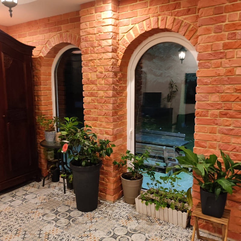 Aranżacja wnętrza z ceglaną ścianą i łukowymi oknami, uzupełniona roślinami doniczkowymi i wzorzystą podłogą. Widoczna część antycznej szafy.