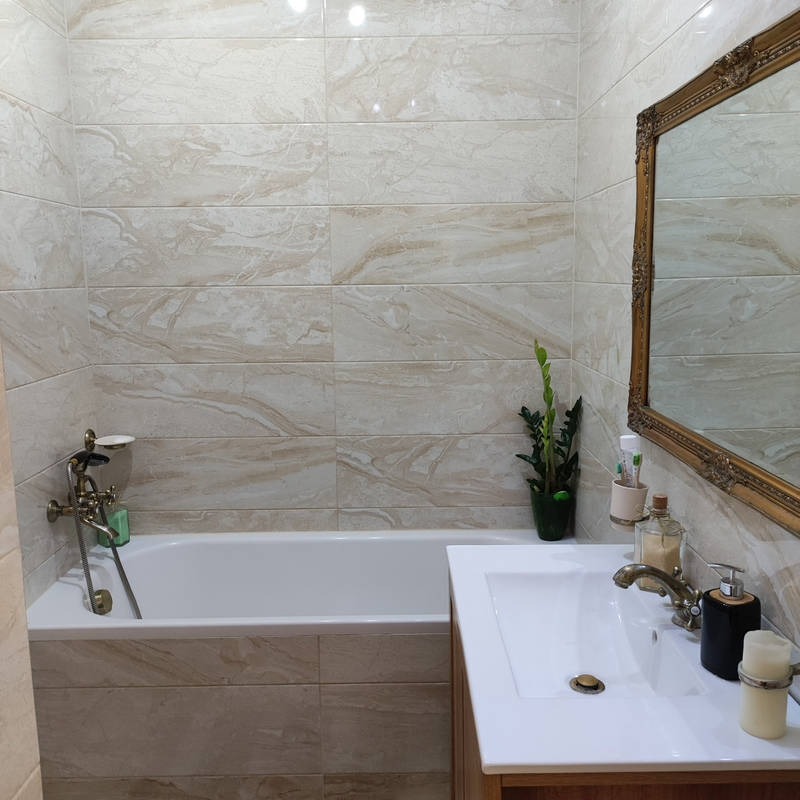 Jasna łazienka z beżowymi płytkami imitującymi marmur, białą wanną i umywalką. Złote detale armatury i ramy lustra dodają elegancji. Roślina w rogu ożywia wnętrze.