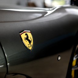 Agencja Marketingowa APOV - Szary samochód sportowy z żółtym logo Ferrari na błotniku. Widoczne detale karoserii i fragment felgi. Oświetlenie podkreśla kształty auta.