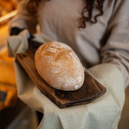 Agencja Marketingowa APOV - Osoba trzyma na desce chleb na lnianej ściereczce. Ciepłe światło podkreśla teksturę skórki. Ujęcie z góry, widoczne dłonie w rękawicach.
