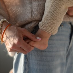 Agencja Marketingowa APOV - Delikatne ujęcie dłoni kobiety w bransoletkach, trzymającej rączkę dziecka w beżowym sweterku, na tle jasnych jeansów. Ciepłe, naturalne światło.