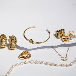 Agencja Marketingowa APOV - Zestaw złotej biżuterii: kolczyki, bransoletka z węzłem, pierścionki (w tym jeden z napisem ZUMA), naszyjnik z perłami i koniczynką, na białym tle z fakturą.