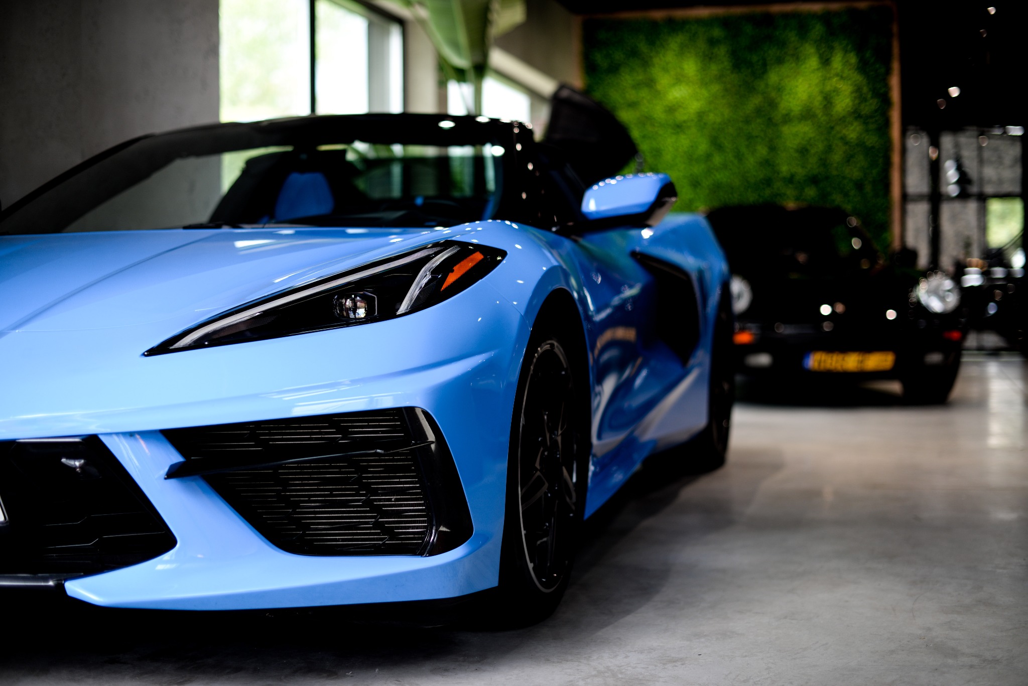 Błękitny Chevrolet Corvette C8 z otwartymi drzwiami na tle zielonej ściany i innego, ciemnego samochodu. Ujęcie z niskiej perspektywy, podkreślające detale przedniego zderzaka.