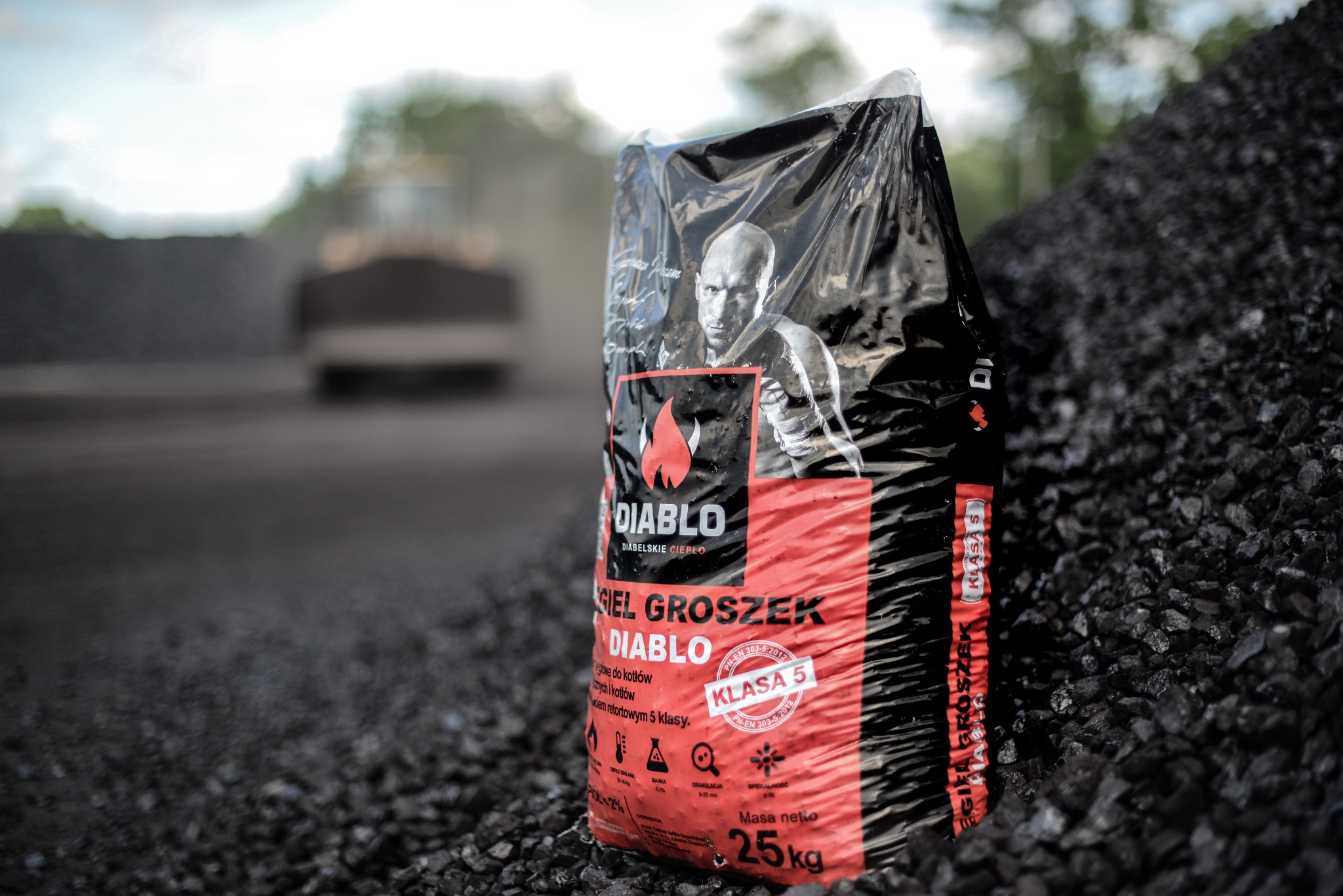 Worek ekogroszku Diablo klasy 5 na tle hałdy węgla i rozmytej sylwetki koparki. Produkt w czarno-czerwonej folii z wizerunkiem osoby. Masa netto 25 kg.