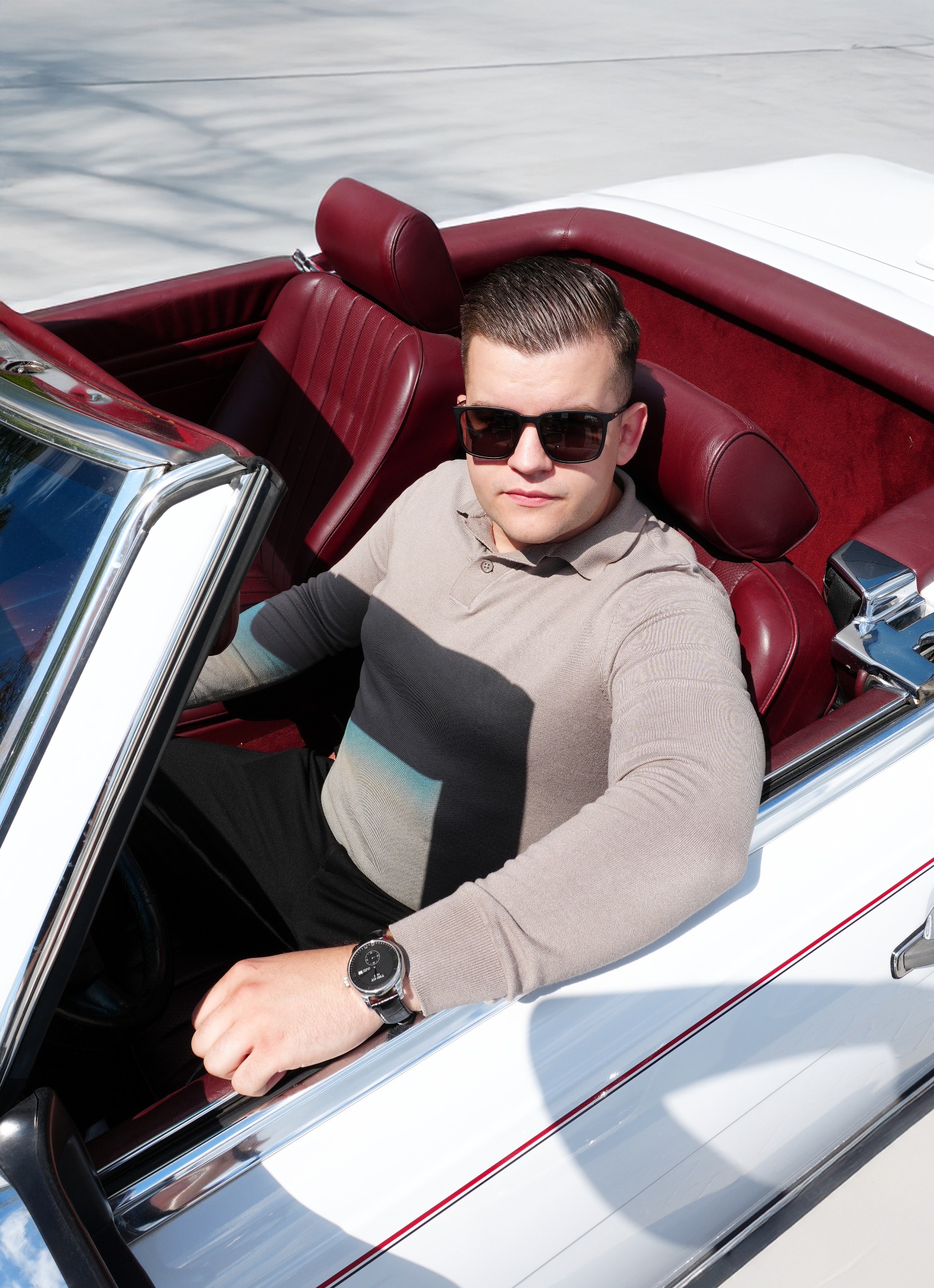 Mężczyzna w okularach przeciwsłonecznych siedzi w białym kabriolecie z bordową skórzaną tapicerką. Widoczny zegarek na ręce i elegancki sweter.