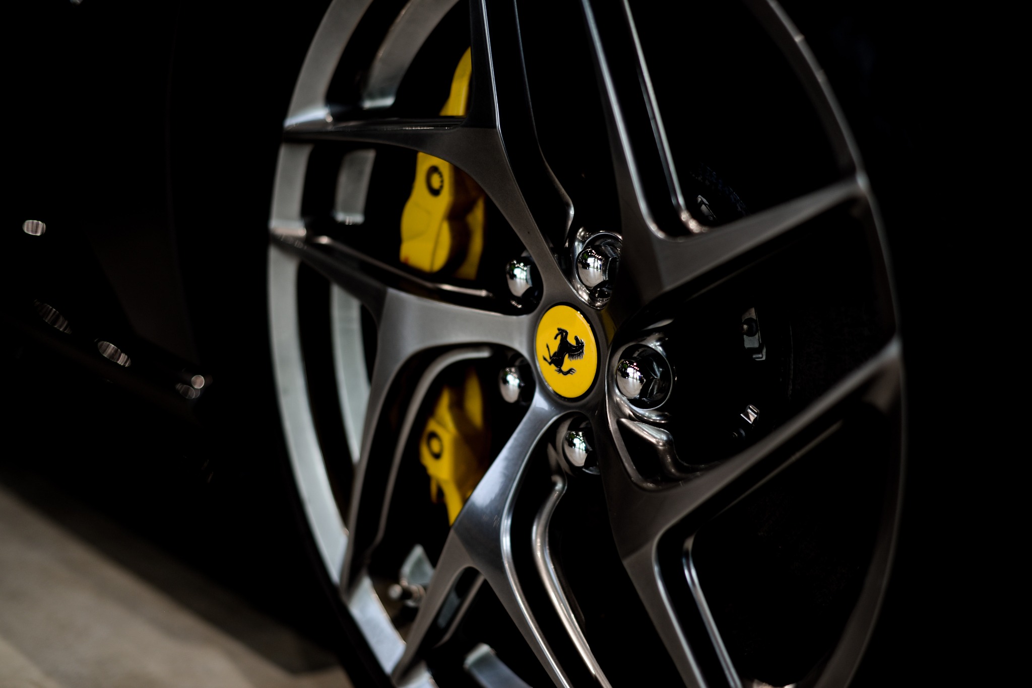 Felga aluminiowa z żółtym zaciskiem hamulcowym i logo Ferrari na tle czarnego lakieru samochodu, uchwycona w zbliżeniu z niskim oświetleniem.