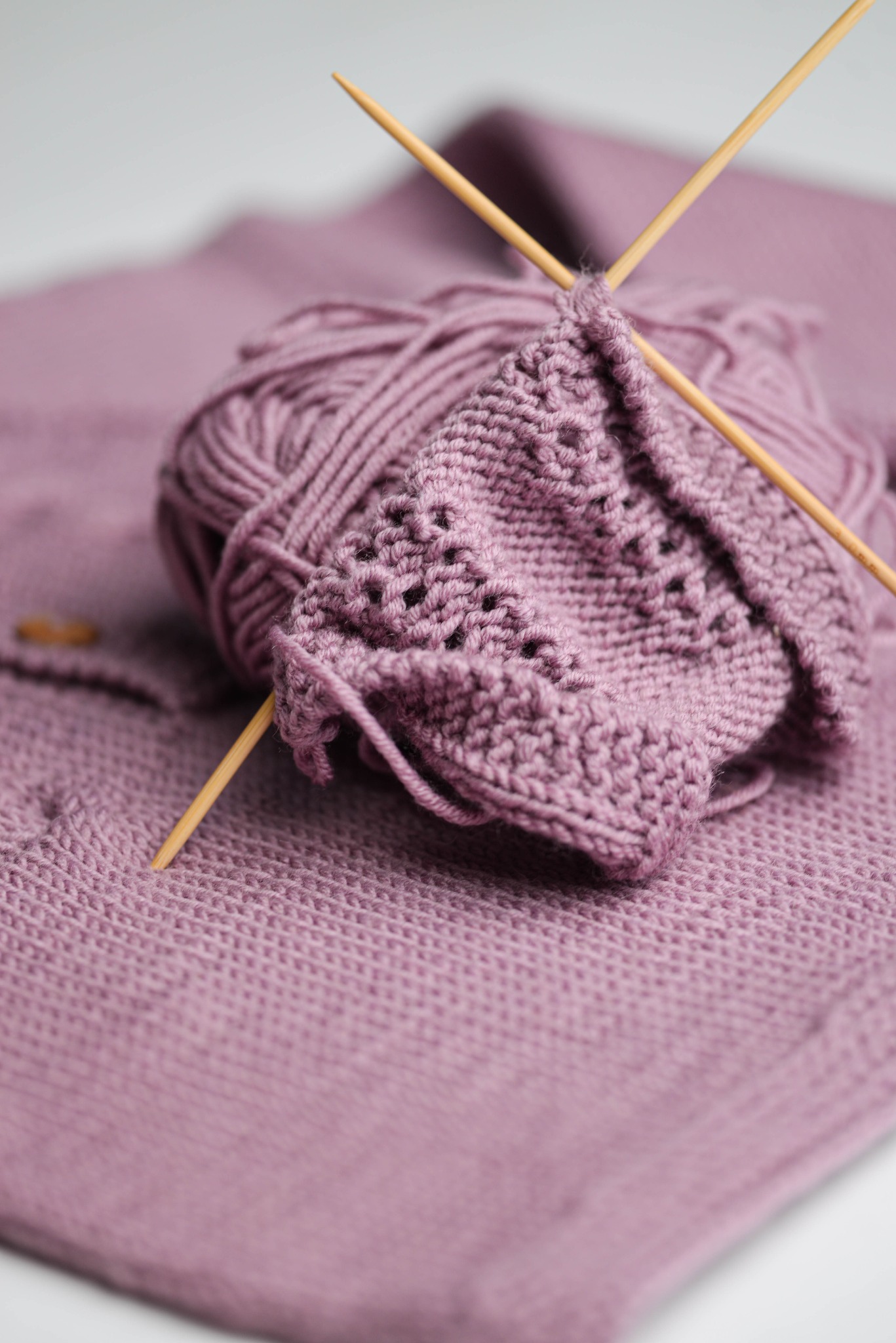 Fioletowa włóczka z drutami na tle dzianiny w odcieniu lila, prezentująca etap tworzenia ażurowego swetra.