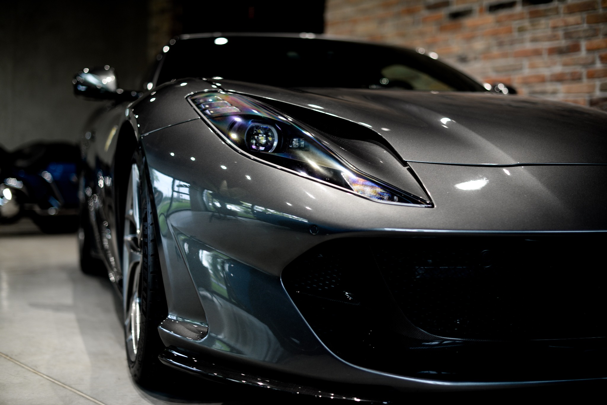 Szary sportowy samochód Ferrari z bliska, z widocznym reflektorem i fragmentem karoserii, na tle ceglanej ściany. Oświetlenie studyjne podkreśla detale.