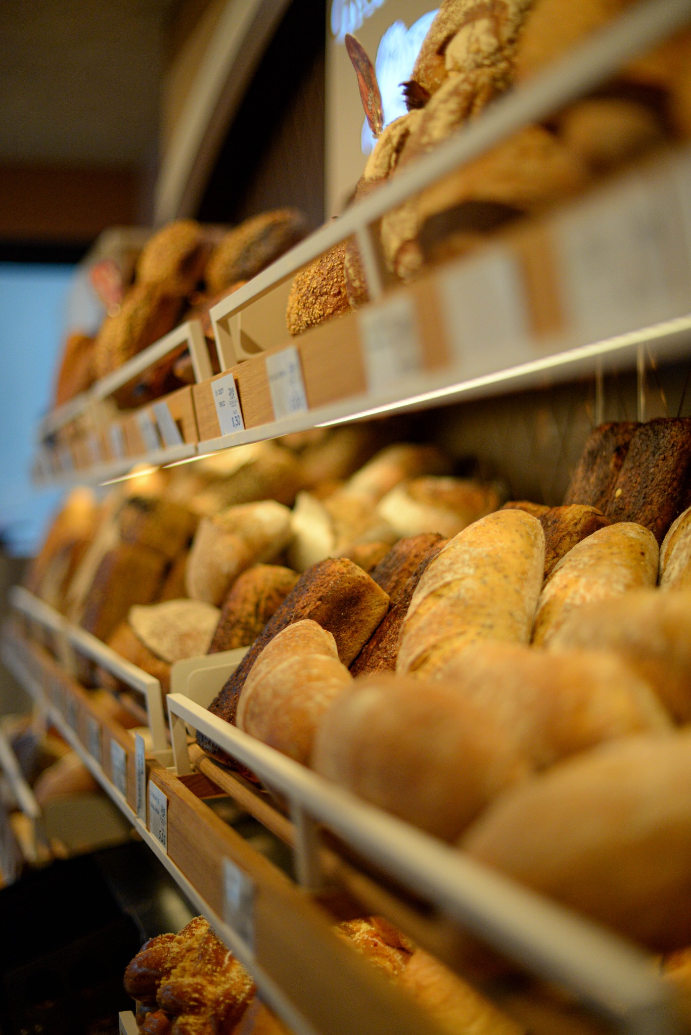 Półki piekarni wypełnione różnorodnym pieczywem: chleby pszenne, razowe i bułki. Widoczne etykiety cenowe. Ciepłe oświetlenie podkreśla świeżość produktów.