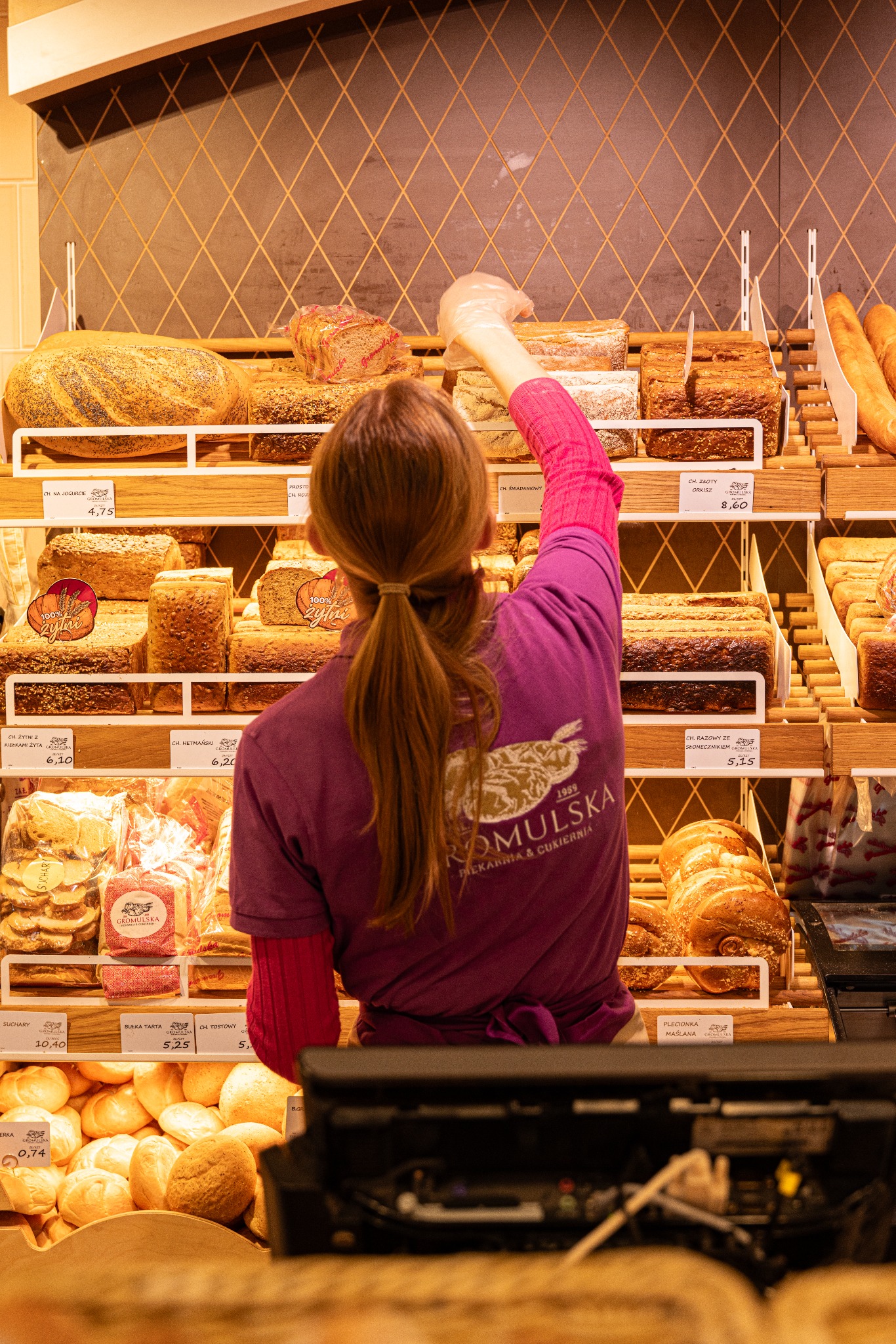 Sprzedawczyni w fioletowej koszulce z logo piekarni Gromulska sięga po pieczywo na półce. Widoczne różne rodzaje chleba i bułek z cenami.