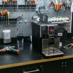 Coffee Cona Marcin Ostrówka - Czarny ekspres do kawy Saeco na warsztatowym stole z narzędziami, sugerujący serwis lub naprawę. W tle perforowana ściana z narzędziami.
