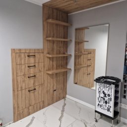 Usługi Budowlane Damian Borys - Drewniana zabudowa w salonie fryzjerskim z lustrem i wózkiem na narzędzia. Biała marmurowa podłoga. Ujęcie z niskiego kąta, podkreślające wysokość zabudowy.