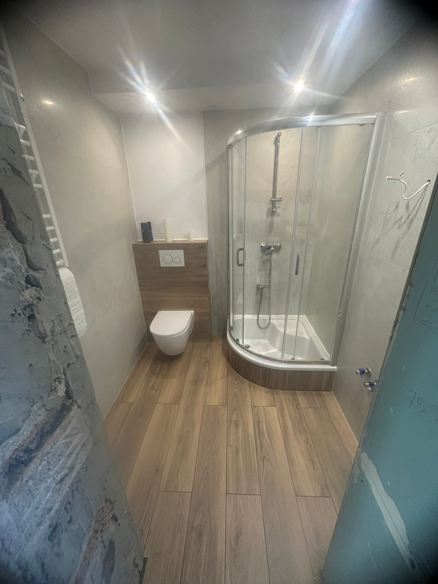 Wykończona łazienka z kabiną prysznicową, podwieszaną toaletą i drewnopodobną podłogą. Ściany w odcieniach szarości. Fragment ściany w stanie surowym po lewej.
