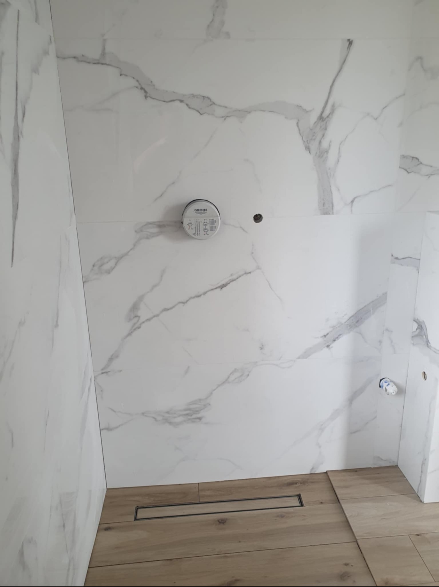 Wykończona łazienka z białymi płytkami imitującymi marmur i drewnopodobną podłogą z odpływem liniowym. Widoczne przygotowane punkty instalacyjne.