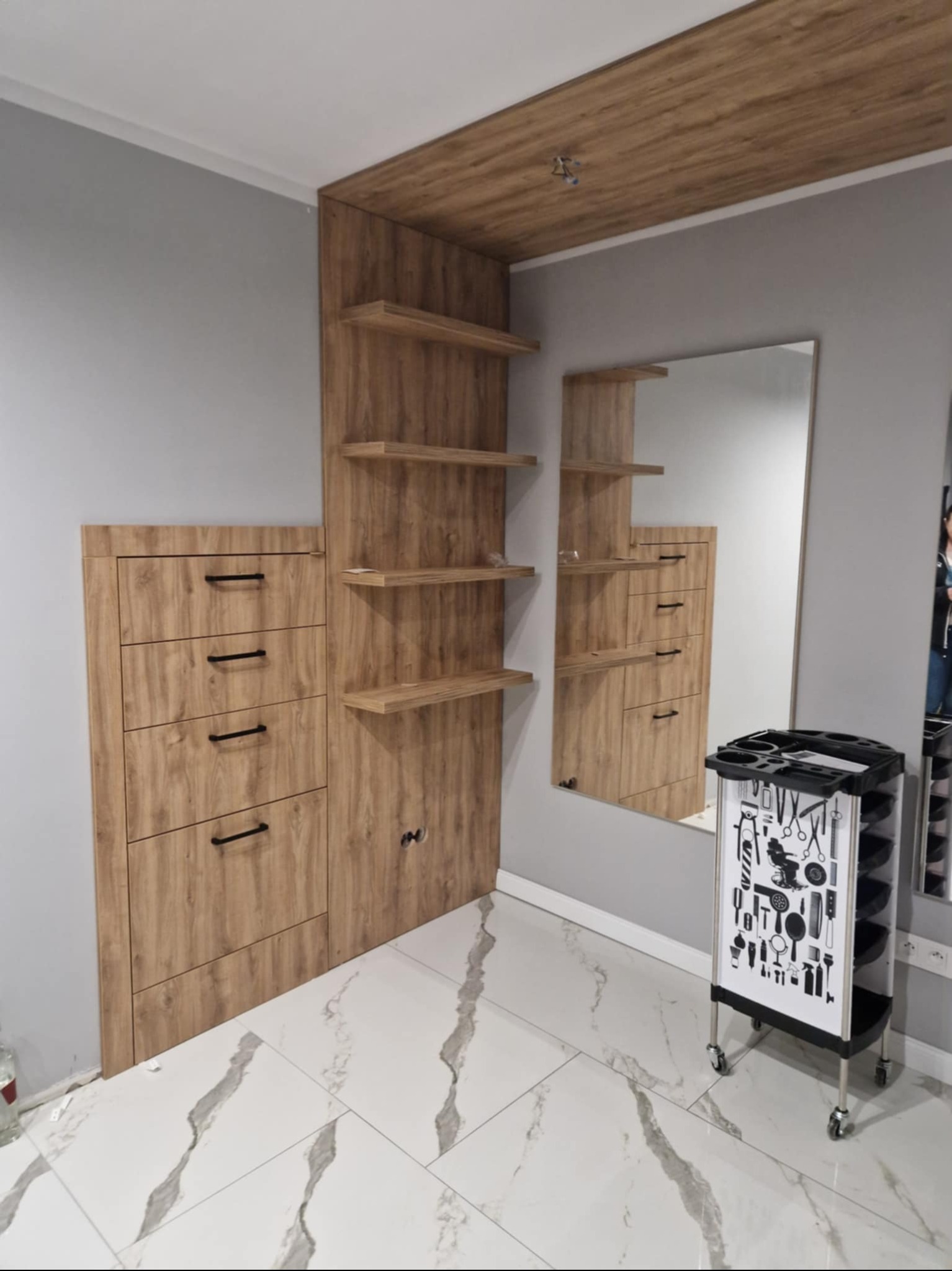 Drewniana zabudowa w salonie fryzjerskim z lustrem i wózkiem na narzędzia. Biała marmurowa podłoga. Ujęcie z niskiego kąta, podkreślające wysokość zabudowy.