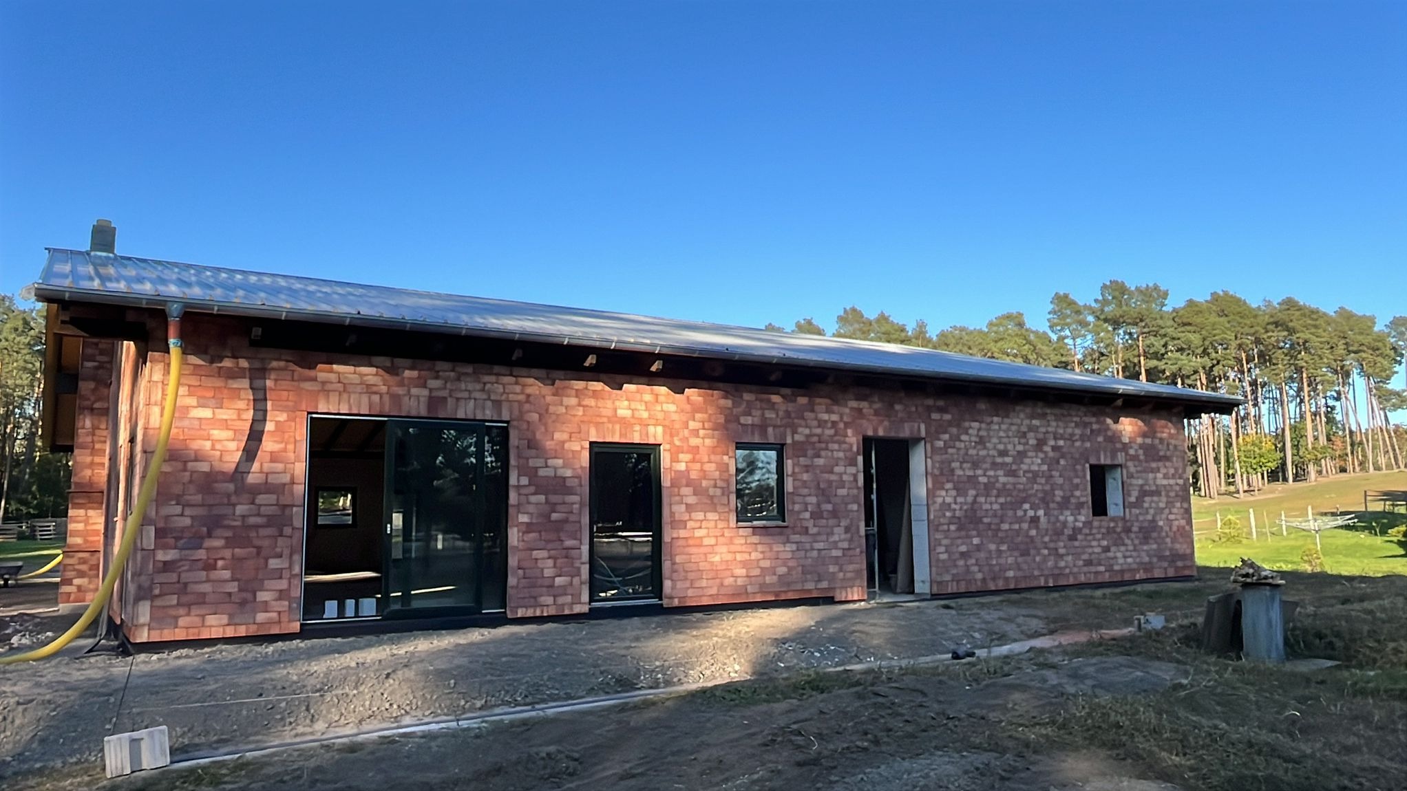 Budynek z cegły klinkierowej w trakcie budowy, widoczne okna i drzwi, teren wokół w stanie surowym, na tle lasu i błękitnego nieba. Ujęcie z zewnątrz, w słoneczny dzień.