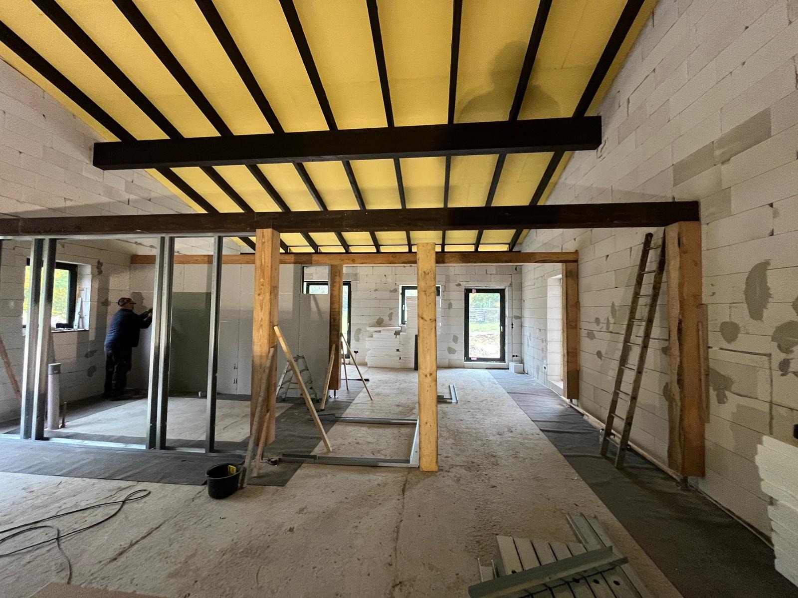 Wnętrze w trakcie remontu: surowy beton, ściany z białych bloczków, drewniane i metalowe konstrukcje, widoczna izolacja sufitu. Pracownik montuje płytę gipsową.