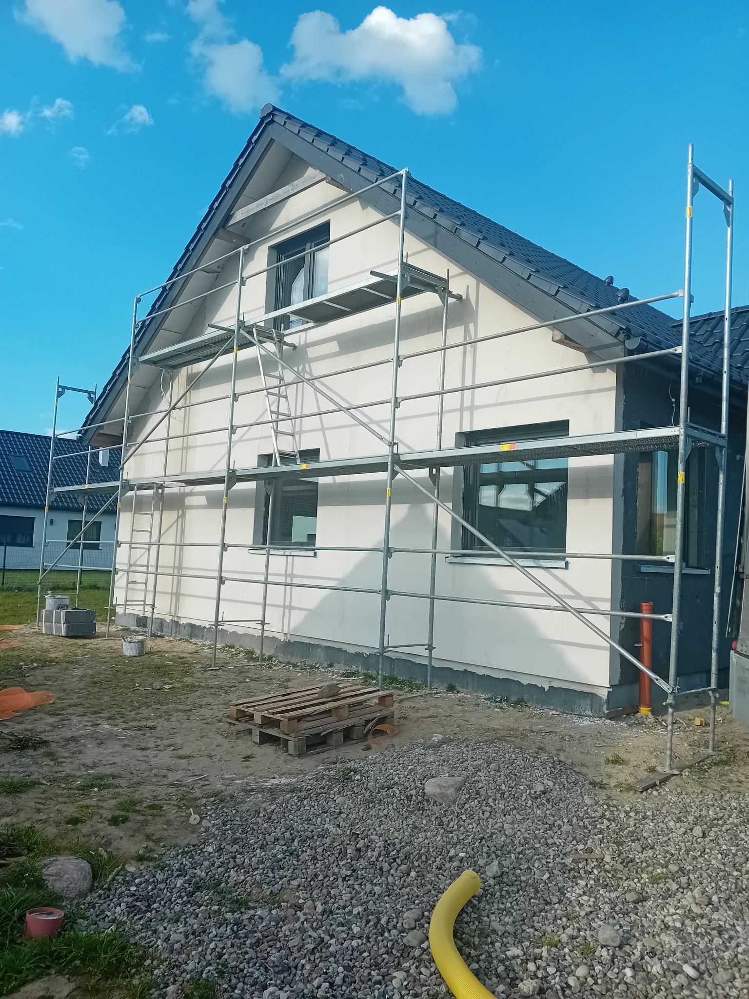 Dom jednorodzinny w trakcie prac wykończeniowych elewacji, otoczony rusztowaniem. Widoczne okna, dach z ciemnej dachówki i fragment niezagospodarowanego terenu.