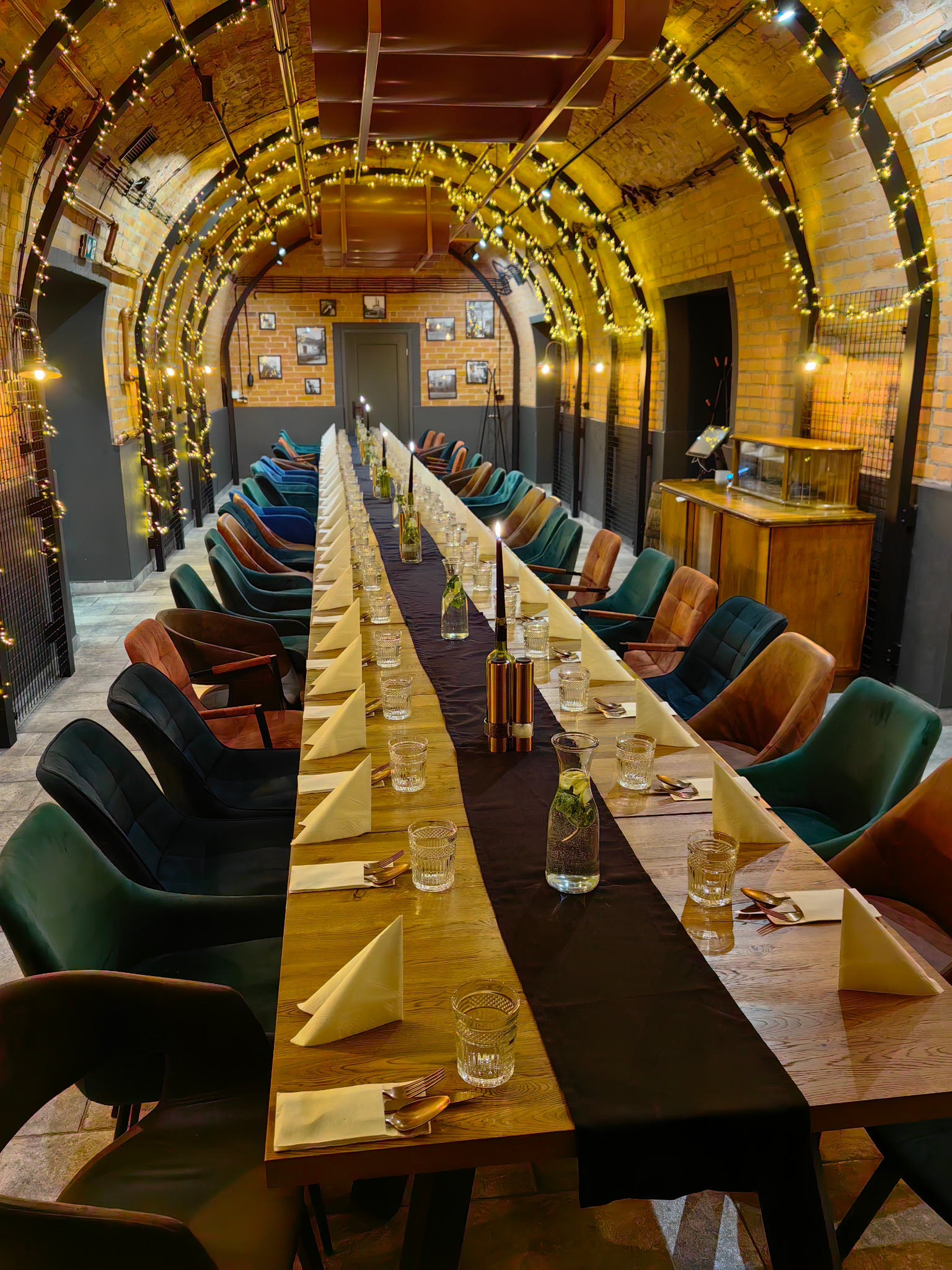 Długi stół z nakryciami i dekoracjami w ceglanym wnętrzu z łukowym sufitem, oświetlonym girlandami. Krzesła w różnych kolorach dodają unikalnego charakteru.