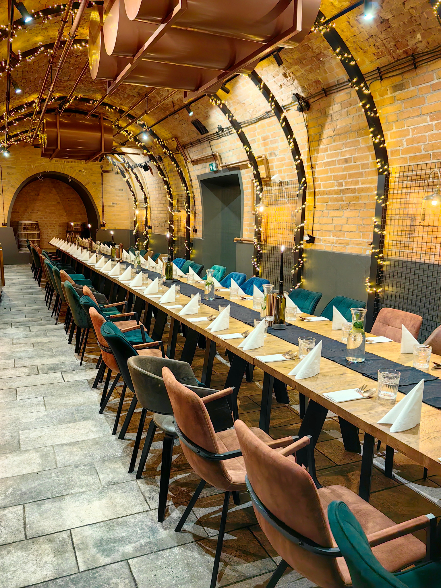Uroczysta aranżacja długiego stołu w ceglanym wnętrzu z łukowym sufitem, oświetlonym girlandami. Przygotowanie do eventu w Łodzi. Różnokolorowe krzesła.