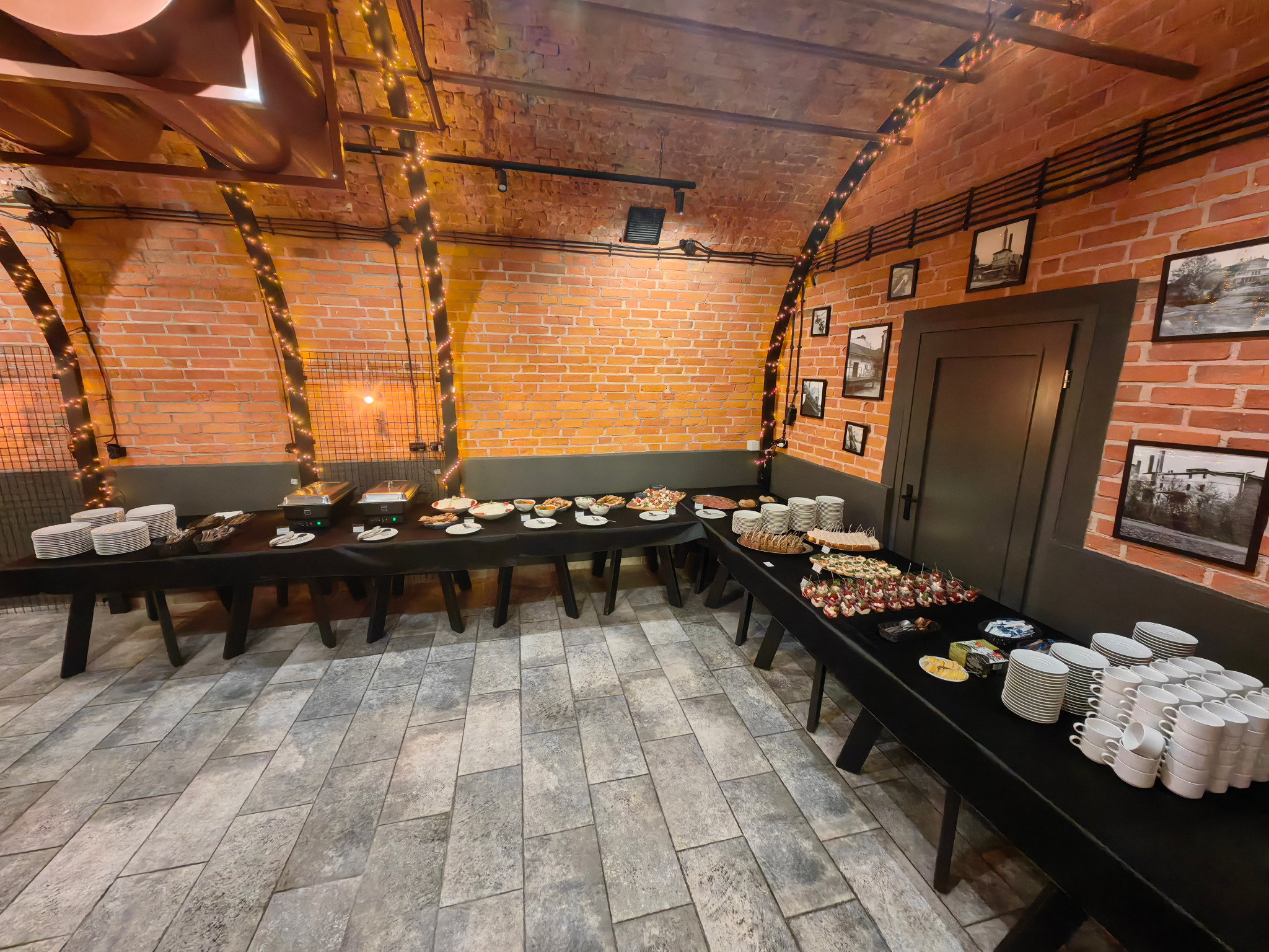 Stół cateringowy z przekąskami i zastawą w industrialnej przestrzeni z ceglanymi ścianami i dekoracyjnym oświetleniem. Widok z perspektywy gościa.