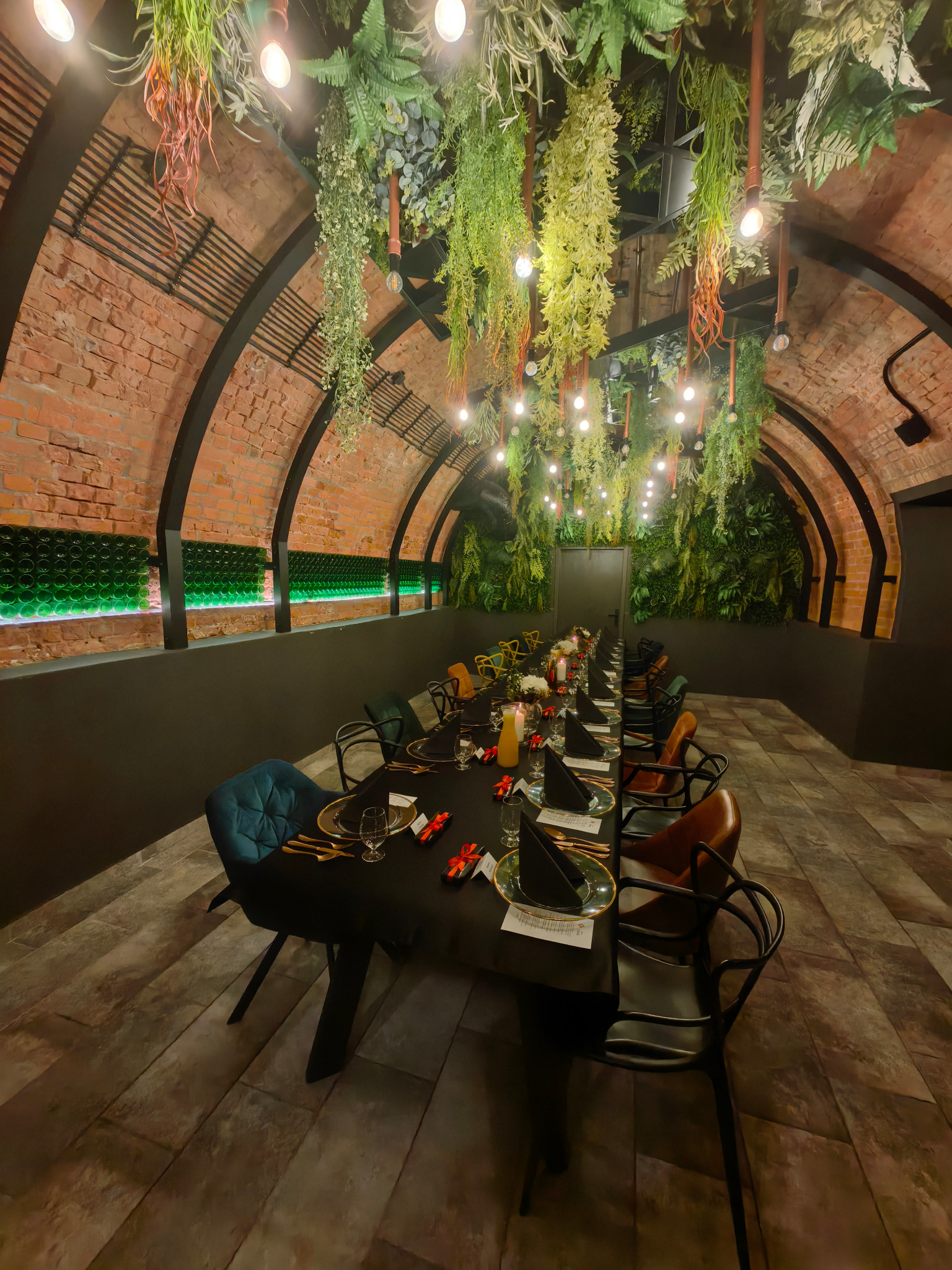 Uroczysta aranżacja długiego stołu w ceglanym wnętrzu z roślinną dekoracją sufitu i butelkową ścianą. Nakrycia, świece i prezenty tworzą elegancki klimat.
