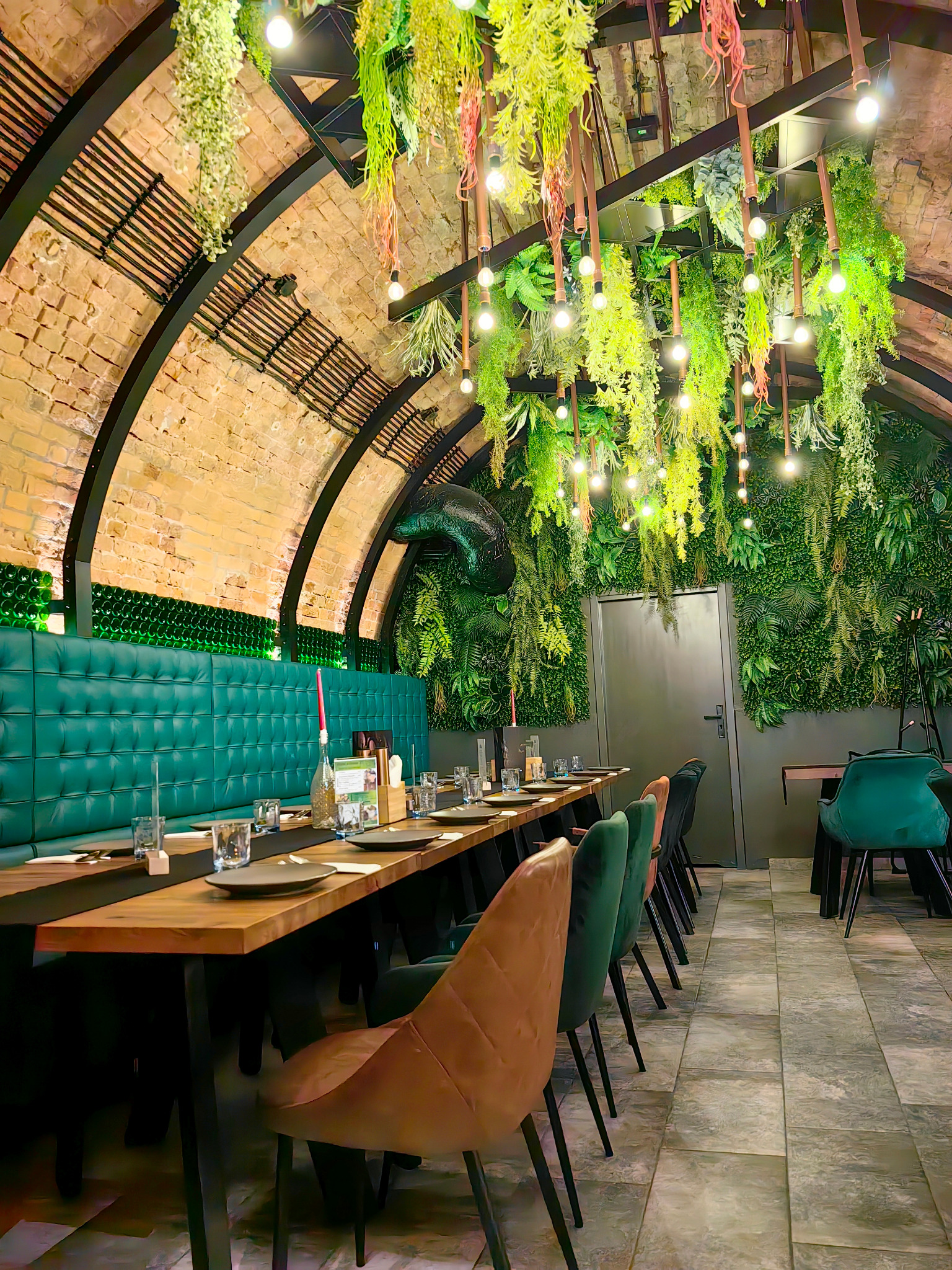 Uroczysta aranżacja stołu w restauracji z ceglanym sklepieniem, zielonymi dekoracjami roślinnymi i eleganckimi krzesłami. Przygotowane nakrycia i oświetlenie tworzą wyjątkowy klimat.
