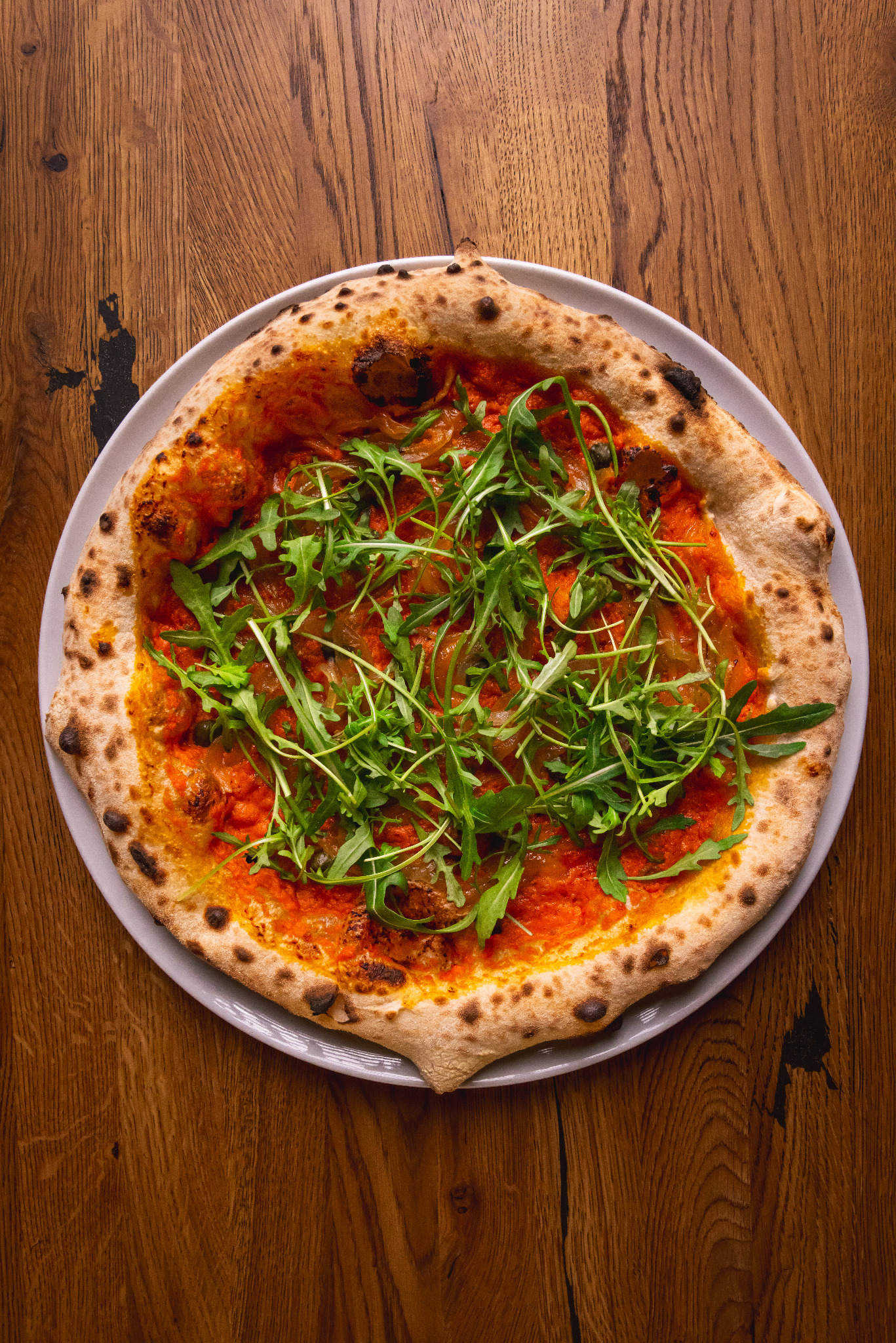 Apetyczna pizza z rukolą na talerzu, prezentowana na drewnianym stole. Widok z góry, podkreślający świeżość składników i rustykalny charakter dania.