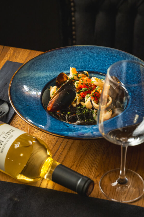 Elegancko podane danie z owocami morza na ciemnym makaronie w niebieskim talerzu, obok butelka białego wina i kieliszek na drewnianym stole.