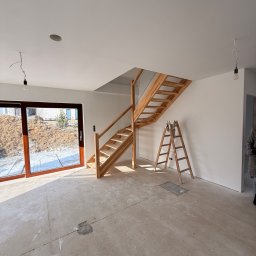 Maciej Rusinek - Wnętrze domu w trakcie wykańczania: drewniane schody, drabina oparta o ścianę, surowy beton na podłodze, ściany przygotowane do malowania, duże okno z widokiem na zewnątrz.