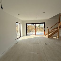 Maciej Rusinek - Jasne wnętrze domu w stanie deweloperskim z drewnianymi schodami i dużymi oknami przesuwnymi. Ściany pomalowane na biało, surowa podłoga betonowa.