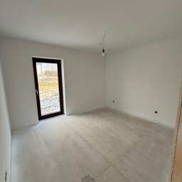 Maciej Rusinek - Jasne wnętrze po szpachlowaniu, z widoczną surową podłogą i świeżo wygładzonymi ścianami. Okno z ciemną ramą wpuszcza naturalne światło.