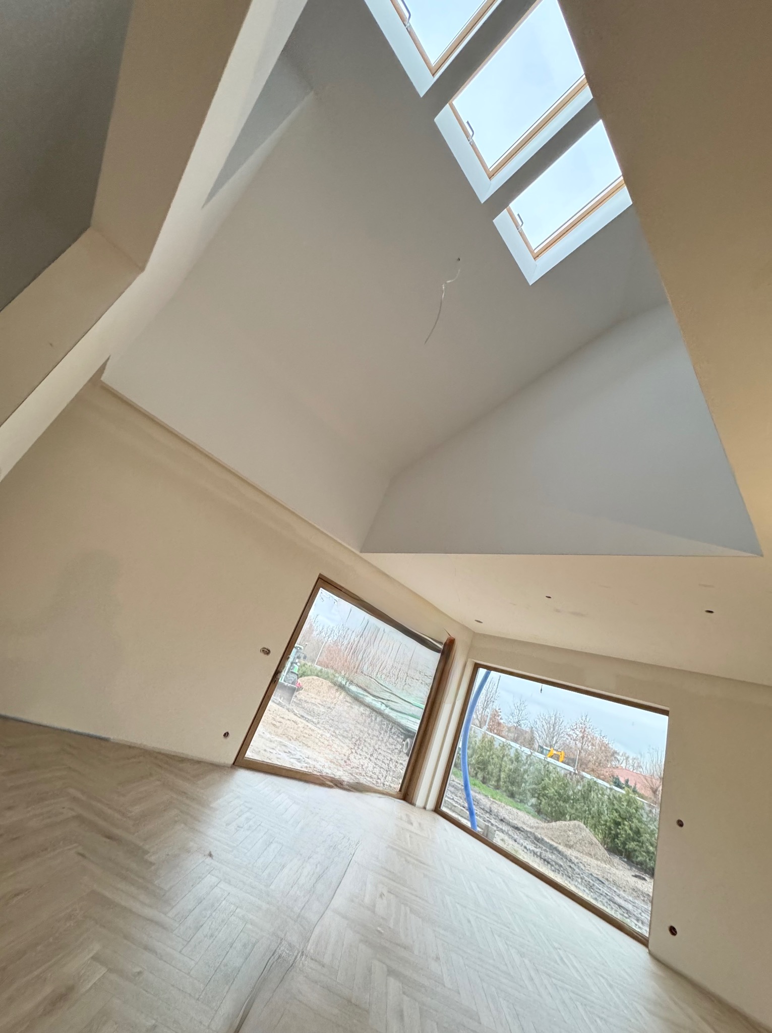 Jasne wnętrze po gładziowaniu ścian i sufitu, z oknami dachowymi i parkietem ułożonym w jodełkę. Widoczne ślady po szpachlowaniu. Dwa okna z widokiem na zewnątrz.