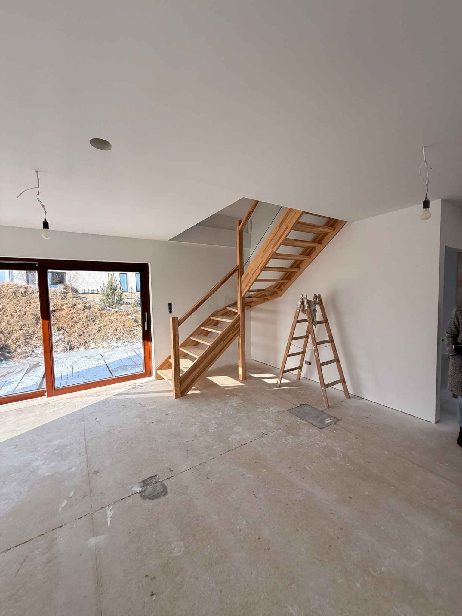 Wnętrze domu w trakcie wykańczania: drewniane schody, drabina oparta o ścianę, surowy beton na podłodze, ściany przygotowane do malowania, duże okno z widokiem na zewnątrz.