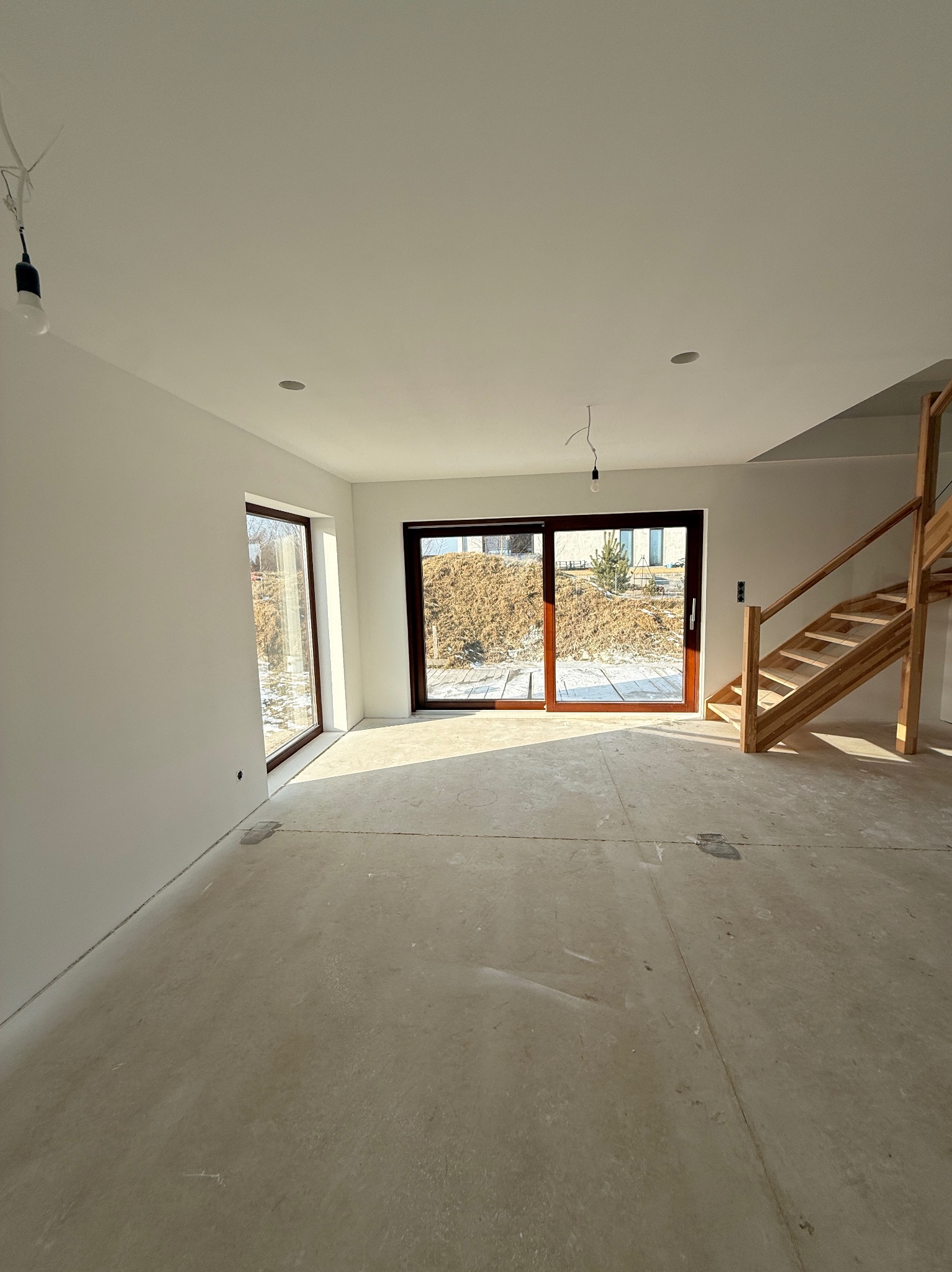 Jasne wnętrze domu w stanie deweloperskim z drewnianymi schodami i dużymi oknami przesuwnymi. Ściany pomalowane na biało, surowa podłoga betonowa.