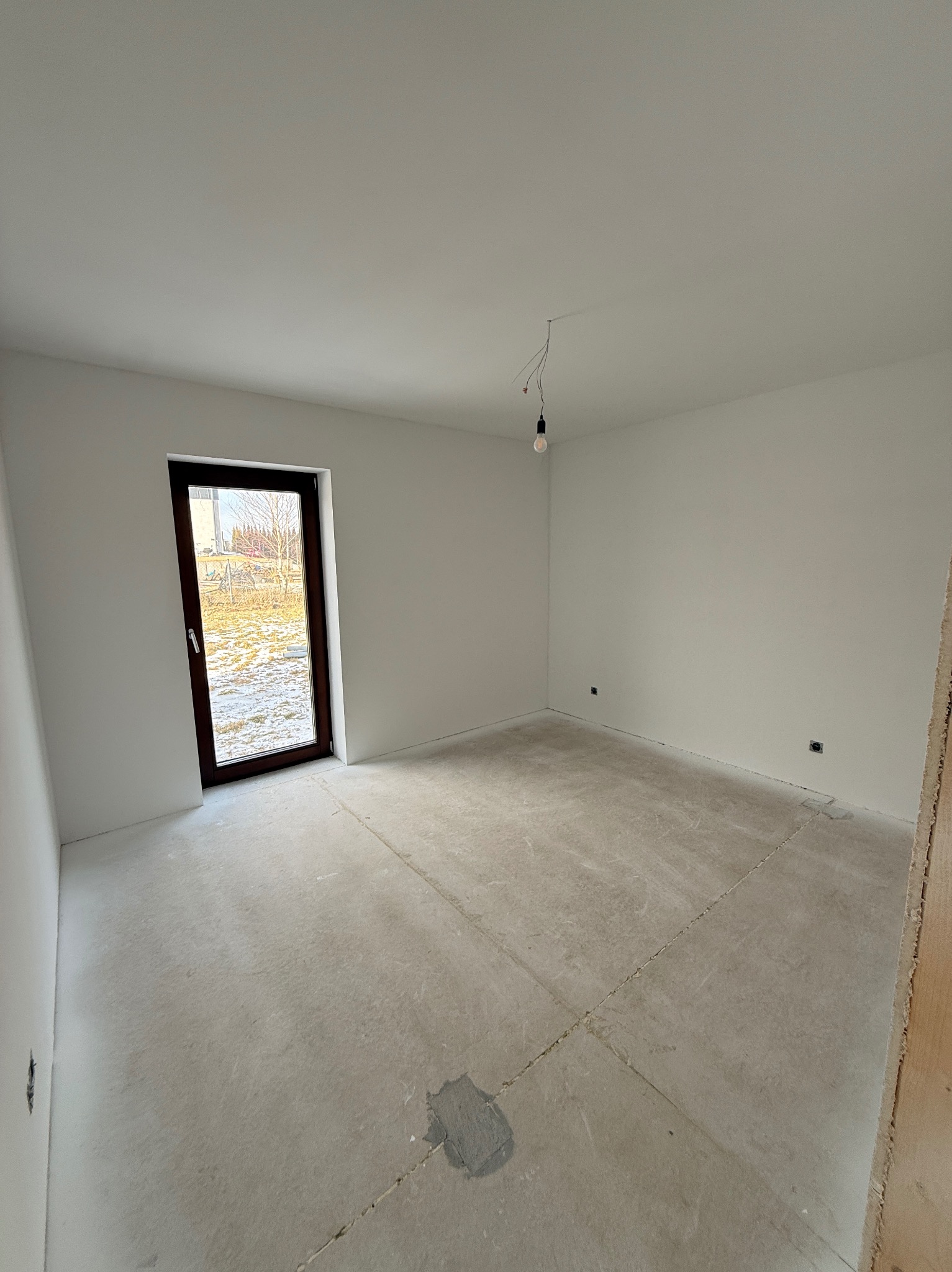 Jasne wnętrze po szpachlowaniu, z widoczną surową podłogą i świeżo wygładzonymi ścianami. Okno z ciemną ramą wpuszcza naturalne światło.