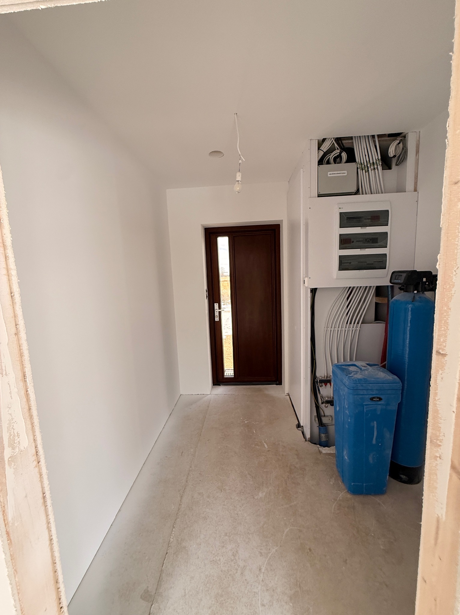 Wnętrze w trakcie wykańczania: białe ściany, brązowe drzwi, instalacja elektryczna i wodna. Surowy beton na podłodze. Widok z perspektywy wejścia.