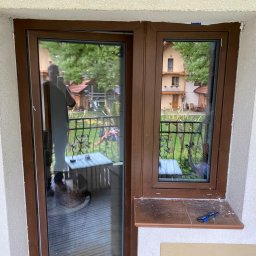 WINFOX GROUP - Brązowe okno balkonowe z uchylnym skrzydłem i małe okno obok, widok na balkon i ogród. Na parapecie leży nóż monterski. W szybie odbija się osoba.