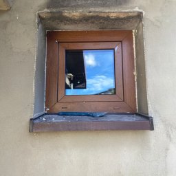 WINFOX GROUP - Montaż okna z brązową ramą w surowej elewacji. Widoczna szpachelka na parapecie i siatka elewacyjna. Okno odbija błękitne niebo z chmurami.